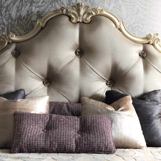 The Romantic Button Upholstered Bed, adorned with an elegant tufted headboard featuring elaborate detailing and six buttons, stands out beautifully against a patterned wallpaper. The bed is tastefully furnished with plush pillows in diverse textures and colors, including metallic and dark shades, bringing a touch of luxury to the bedroom decor.
