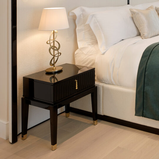 An Art Deco Style Bedside Table with a black glossy finish and gold accents, adorned with a decorative table lamp featuring an intricate gold base and white shade. The nightstand is situated next to a neatly made bed with white bedding and a green throw.