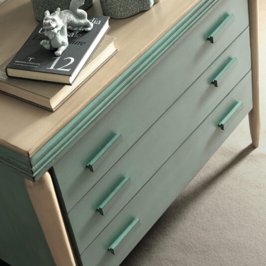 Luxury Art Deco Style Chest Of Drawers A close-up of the Luxury Art Deco Style Chest Of Drawers, a green wooden dresser with three drawers featuring horizontal handles. On top are a closed book with a dolphin figurine, another book, and a decorative ceramic item. The dresser stands on a beige carpeted floor.
