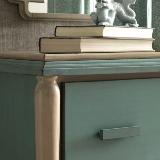 Luxury Art Deco Style Chest Of Drawers A close-up of the green Luxury Art Deco Style Chest Of Drawers with a beige handle, featuring a stack of three books and a small decorative dragon figurine on top. Behind the chest is a textured wall with an angular-framed mirror partially visible.