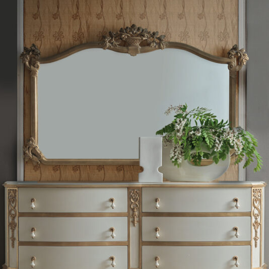 A vintage wooden dresser with ornate gold details is highlighted by a Large Rococo Style Overmantle Mirror. On top of the dresser, a modern white vase filled with green foliage and small white flowers enhances the textured wallpaper backdrop.
