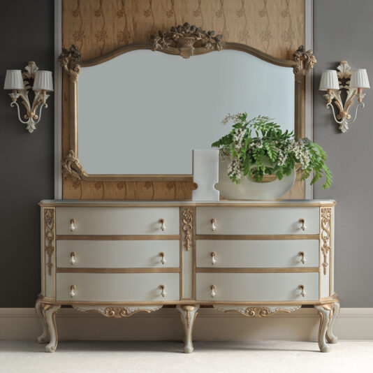 Large Rococo Style Overmantle Mirror An elegant dresser showcases a Large Rococo Style Overmantle Mirror, accompanied by two wall sconces. A vase containing green foliage and white flowers rests on the dresser, bringing a fresh element to the classic decor.
