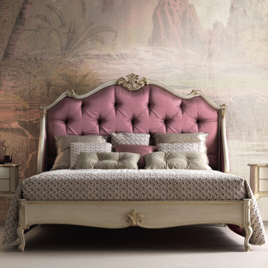 A vintage-style bedroom showcases a plush, pink, tufted headboard with intricate carvings at the top, reminiscent of the Classic Louis Style Button Upholstered Bed (Copy). The bed is dressed with patterned bedding and is accented by numerous decorative pillows. The walls feature a soft, artistic mural. Cream-colored bedside tables stand on either side of the bed.