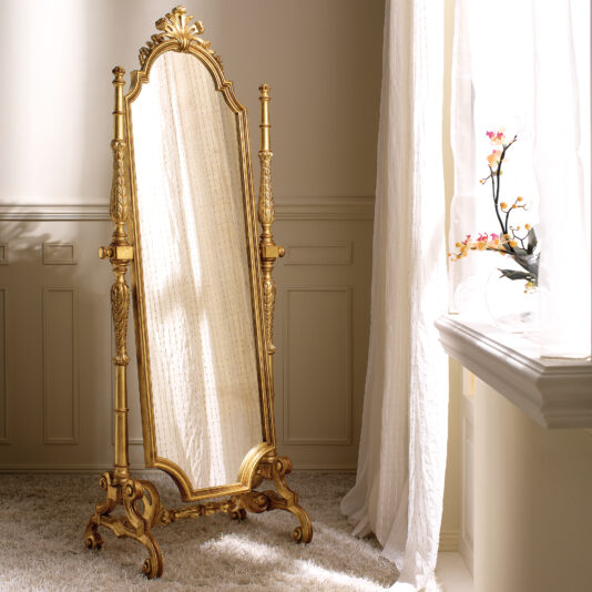 A vintage Freestanding Rococo Style Tilting Mirror with ornate gold carvings stands elegantly on a plush white carpet. Sunlight filters through a window draped with sheer curtains, creating gentle shadows. A vase filled with delicate flowers adorns the windowsill.