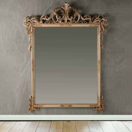 The Large Baroque Style Wall Mirror, distinguished by its intricate, decorative frame with elaborate scrollwork and floral designs, is mounted on a textured gray wall. Part of a wooden floor can be seen at the bottom.