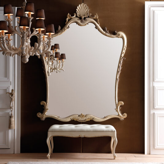 The elegant room features a Large Rococo Style Wall Mirror with a decorative frame. Below the mirror is a cushioned bench with elegantly curved legs. On the left, a classic chandelier adorned with black lampshades hangs, offering a striking contrast to the brown and white walls.