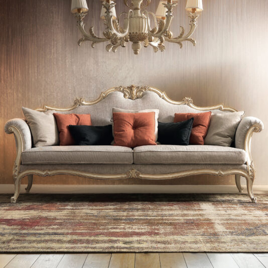 The Ornate Luxury Rococo Style Sofa, featuring intricate carvings and upholstered in beige fabric, is complemented by black and brown cushions. It emanates a sense of luxury as it rests on a patterned rug beneath an elaborate chandelier, set against a textured, warm-colored wall.