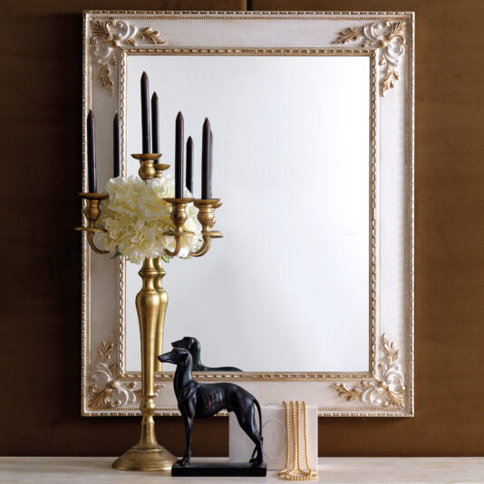 A Rococo Style Rectangular Wall Mirror featuring an ornate frame is complemented by a brass candelabra with black candles and white flowers, accompanied by a small black dog sculpture and a curated stack of elegant books, all artfully displayed on a table against a brown backdrop.