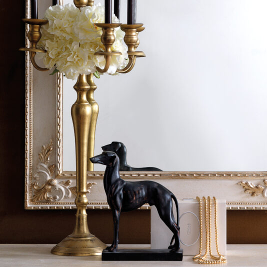 Rococo Style Rectangular Wall Mirror In a decorative setting, the Rococo Style Rectangular Wall Mirror, featuring lavish ornate detailing, is beautifully complemented by a brass candle holder with white flowers. Nearby, a black dog figurine, a small white book, and a string of pearls rest on the light-colored surface.