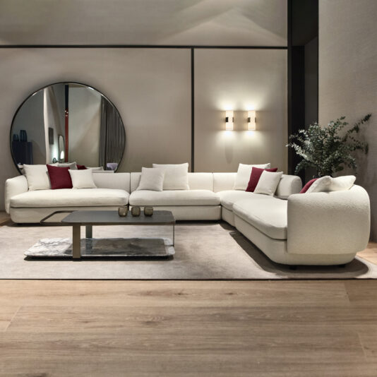 A modern living room showcases a Large Modular Contemporary Corner Sofa in an L-shape, adorned with red and white cushions. Behind this stylish piece, a circular mirror is mounted on the wall. A sleek coffee table rests on a beige rug, complemented by a potted plant that introduces greenery to the space.