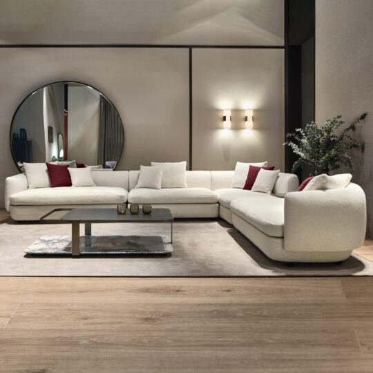 A modern living room showcases a Large Modular Contemporary Corner Sofa in an L-shape, adorned with red and white cushions. Behind this stylish piece, a circular mirror is mounted on the wall. A sleek coffee table rests on a beige rug, complemented by a potted plant that introduces greenery to the space.