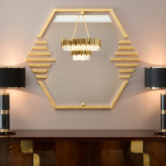 Art Deco Inspired Hexagonal Mirror A modern Art Deco-inspired interior features the Art Deco Inspired Hexagonal Mirror with gold accents on a taupe wall. Reflected is a geometric chandelier above a dark wood console with gold details, flanked by two black and gold lamps, adding elegance and charm. Also featuring high-end furniture in the form of a dark wood cabinet