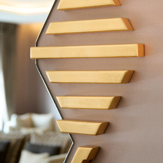 Art Deco Inspired Hexagonal Mirror Close-up of an Art Deco design featuring gold bars on a light brown wall with a softly blurred background showing the reflection in an Art Deco Inspired Hexagonal Mirror, glimpsing neutral furniture and curtains.