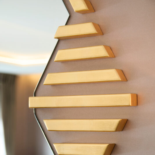 Art Deco Inspired Hexagonal Mirror A close-up view of the Art Deco Inspired Hexagonal Mirror showcases a zigzag pattern with gold-colored bars on a smooth, light brown backdrop, blending geometric elements for a modern and stylish aesthetic.