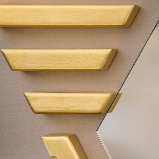 Art Deco Inspired Hexagonal Mirror A close-up of a wall showcases geometric patterns with gold textured blocks in diagonal lines on a beige background. The Art Deco Inspired Hexagonal Mirror is visible on the right, introducing a reflective element to the stylish scene.