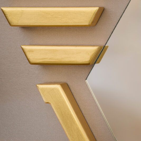 Art Deco Inspired Hexagonal Mirror Close-up of the Art Deco Inspired Hexagonal Mirror partially reflecting a wall, featuring three gold geometric wooden pieces on a light brown textured surface, creating an abstract visual against the muted background.