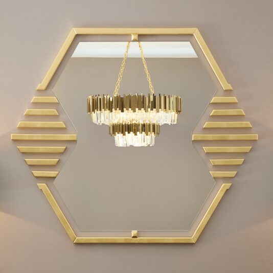 The Art Deco Inspired Hexagonal Mirror, featuring a gold frame and horizontal gold accents, reflects a stylish chandelier adorned with gold metal and crystal pendants, enhancing its elegant modern appeal.