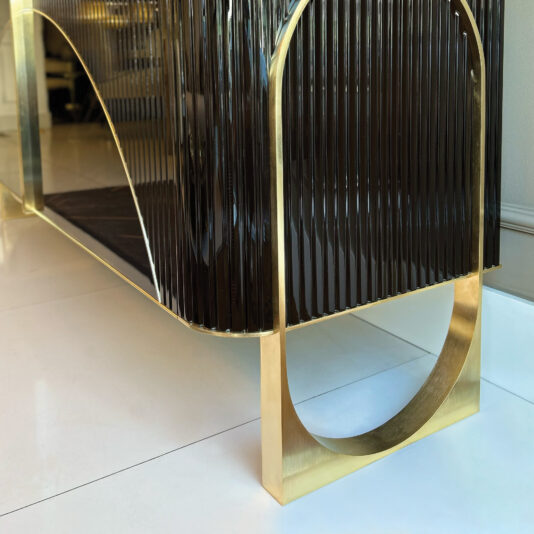 A close-up of the Luxury Art Deco Style Sunburst Sideboard, featuring glossy dark ribbed panels and gold metal legs in a geometric semi-circular design, displayed on a white tiled floor.