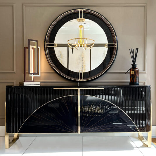 The Luxury Art Deco Style Sunburst Sideboard, finished in glossy black with gold accents, stands against a taupe wall. A large round mirror above reflects a gold chandelier, while decor like a lamp and diffuser adorns the sideboard.