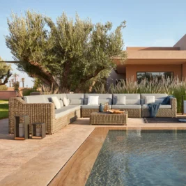 Outdoor patio scene with a large wicker sectional sofa adorned with light gray cushions and blue pillows, perfect for outdoor entertaining. A wicker coffee table holds decorative items. The area is next to a pool, flanked by olive trees, tall grasses, and a beige house in the background.