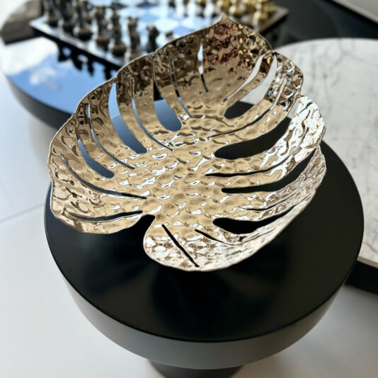 The Tropical Leaf Basket, designed in a metallic monstera leaf shape, rests on a black round table. In the blurred background, there's a marble-topped table and a chess set.