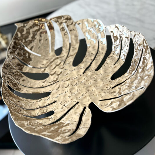 Tropical Leaf Basket The Tropical Leaf Basket, featuring a hammered gold metallic finish and shaped like a large monstera leaf, sits on a dark round table.
