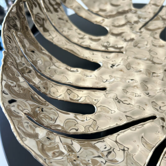 Tropical Leaf Basket A close-up of the Tropical Leaf Basket shows its metallic gold finish, hammered texture, and cutout design resembling a stylized leaf or fan. The shiny surface reflects light for a glossy effect.