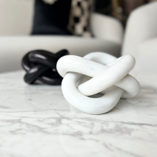 A white ceramic knot sculpture sits on a marble table, with a black knot sculpture blurred behind. Modern white furniture surrounds the space, accented by a Set of 3 Burr Walnut Effect Photo Frames on the wall.