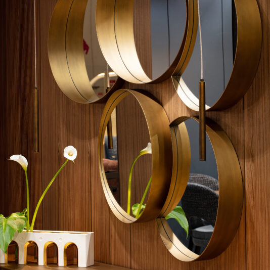 A modern interior showcases the Modern Floating Wall Mirror Cluster with circular gold frames on a wood-paneled wall, accompanied by a white vase with three calla lilies and a small decorative white piece on the wooden surface.