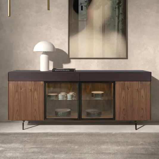 A Modern 4 Door Walnut Veneer Sideboard with wood and glass doors showcases crockery, topped with a white lamp and book, placed against a textured wall beneath abstract artwork.
