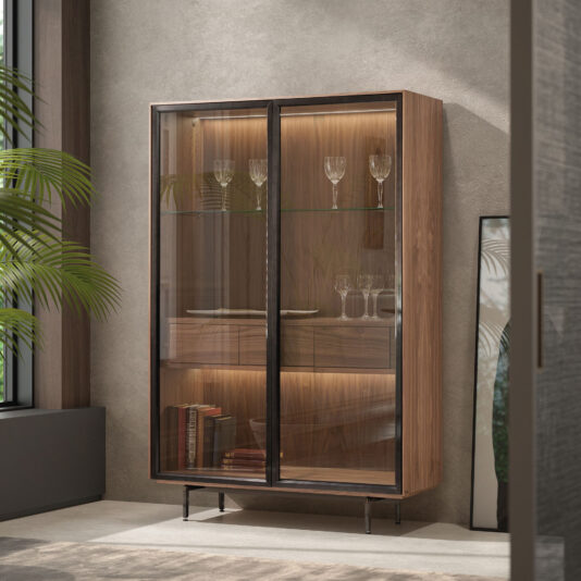 The Modern Walnut Veneer Display Cabinet with glass doors stands against a textured wall, its warm interior lighting illuminating displayed glassware. A nearby leafy plant adds a fresh touch of greenery to the stylish scene.
