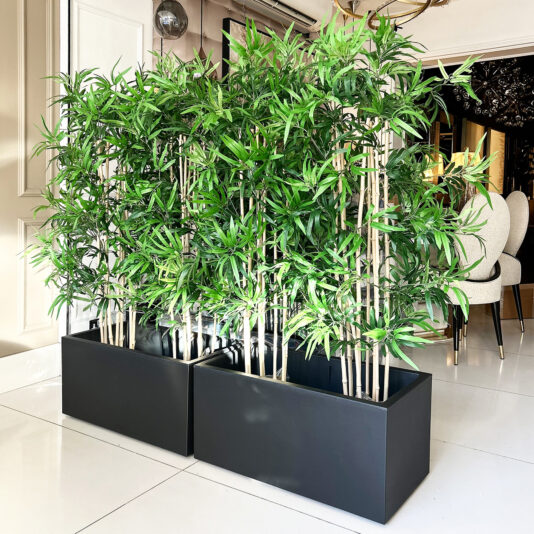 Tall green bamboo in rectangular black planters stand indoors on a shiny white tiled floor. Elegant dining chairs, a chandelier, and the Faux White Agapanthus In Glazed Bowl (Copy) complete the sophisticated space.