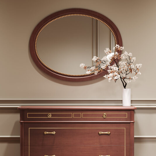 A Classic Style Cherrywood Chest of Drawers (Copy) with gold accents stands against a beige wall, topped with a white vase of flowering branches. Above, an oval mirror with a wooden and gold-trimmed frame reflects the room.