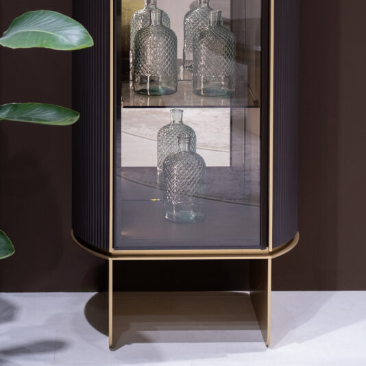 The Contemporary Lacquered Narrow Display Cabinet with gold trim features three textured glass bottles on two shelves. A green plant leaf sits to the left, against a brown wall and light flooring.