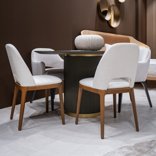 A modern dining area highlights the Round Contemporary Lacquered Dining Table, decorated with a vase and surrounded by four white upholstered chairs with wooden legs, all set against brown walls and stylish décor.
