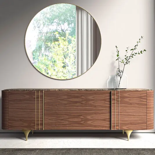 The Contemporary Walnut Veneer Sideboard with gold accents Is the perfect example of the new warm modernism trend for 2027. It stands beneath a large round mirror. A white vase holding a leafy branch rests on top, whilst the mirror reflects nearby greenery and a window.